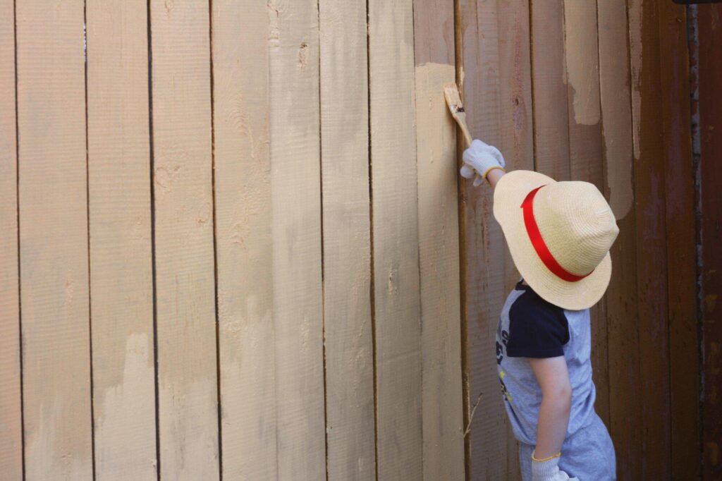 young child wearing sun hat and gloves painting wooden fence